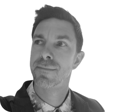 A grayscale photo of a man with light stubble wearing a jacket and a patterned shirt.