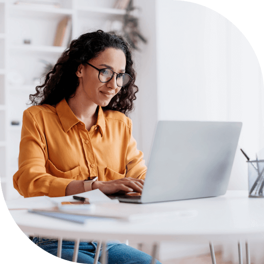 Woman in yellow blouse using laptop at a white desk in a bright office space.