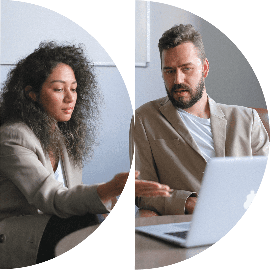 Two professionals in a business meeting, with a woman gesturing towards a laptop and a man listening attentively.