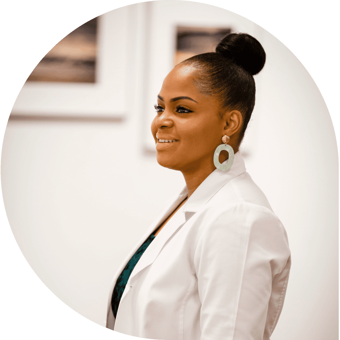 A woman with her hair in a bun, wearing a white blazer and hoop earrings, smiles profile view in an art gallery.