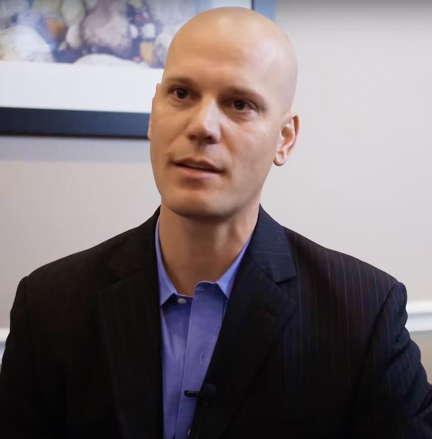 A bald man in a dark suit and blue shirt sitting in an office environment.