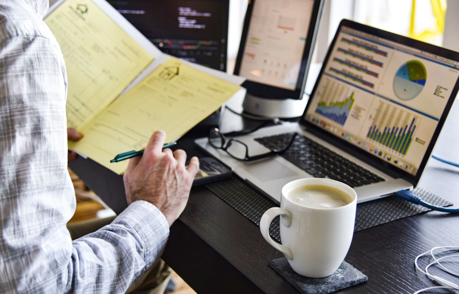 Person in a white shirt reviewing documents at a desk with a laptop, two monitors displaying graphs, and a cup of coffee.
