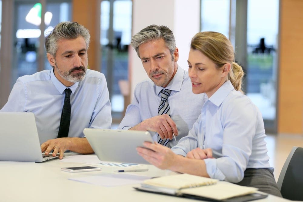 Three business professionals, two men and a woman, are sitting at a table, looking at a tablet while discussing work. There are laptops, papers, and notebooks on the table.